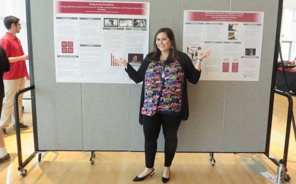 The image shows Davi Kallman standing beside a large research poster mounted on a display board in a hallway or conference setting. She is gesturing toward the poster while speaking, appearing engaged and mid-explanation.