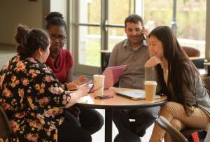 Four colleagues sit around a table in a bright common area, engaged in conversation and collaboration. A laptop, tablet, coffee cups, and notepad are on the table as the group discusses ideas in a welcoming, professional environment.