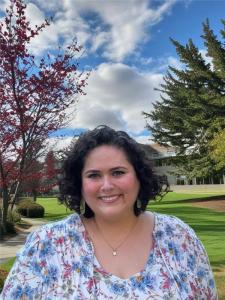 Photo of Davi Kallman. This is a bust portrait of Davi, she is wearing a white and blue floral long sleeve shirt, she has short curly brown hair, and she is standing in front of Pierce College. There are clouds and trees in the background. 