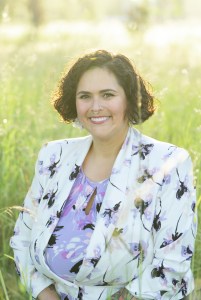 Professional headshot of a smiling person with short, dark curly hair wearing a white blazer with a purple floral print over a lavender floral blouse. They are seated outdoors in a sunlit grassy field with a softly blurred green background, creating a warm, natural portrait.