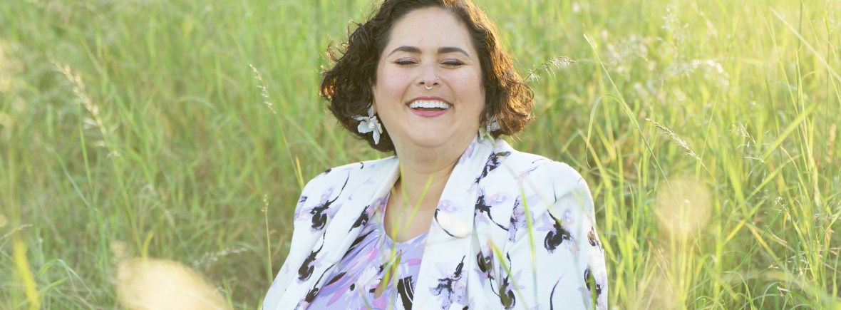 Professional headshot of Dr. Davi Kallman smiling while seated in a sunlit field of tall grass, wearing a floral blazer and purple top, conveying warmth, approachability, and professionalism.