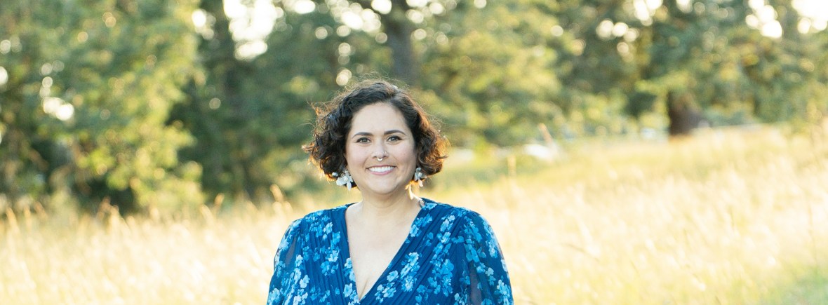 Professional portrait of Dr. Davi Kallman smiling in a sunlit meadow, wearing a blue floral dress with sheer sleeves. Golden grass and softly blurred trees create a warm, natural backdrop that conveys confidence, warmth, and approachability.